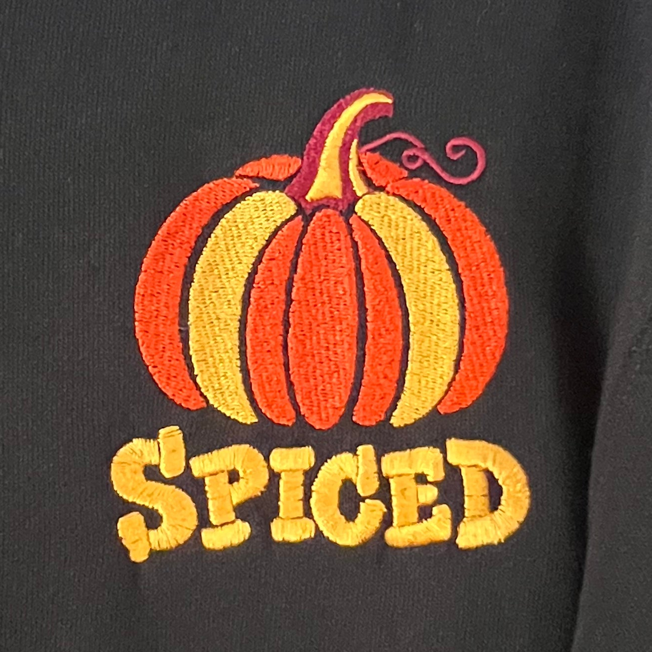 A black zip-up hoodie featuring an embroidered orange and yellow pumpkin graphic above the word 'SPICED' in yellow letters, shown against a light background.